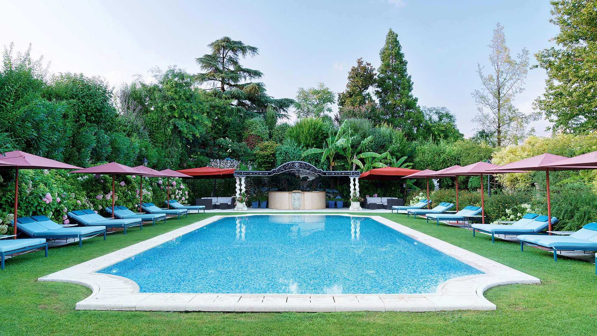 Outdoor pool surrounded by greenery with blue loungers and terracotta umbrellas, a woman walking along the edge