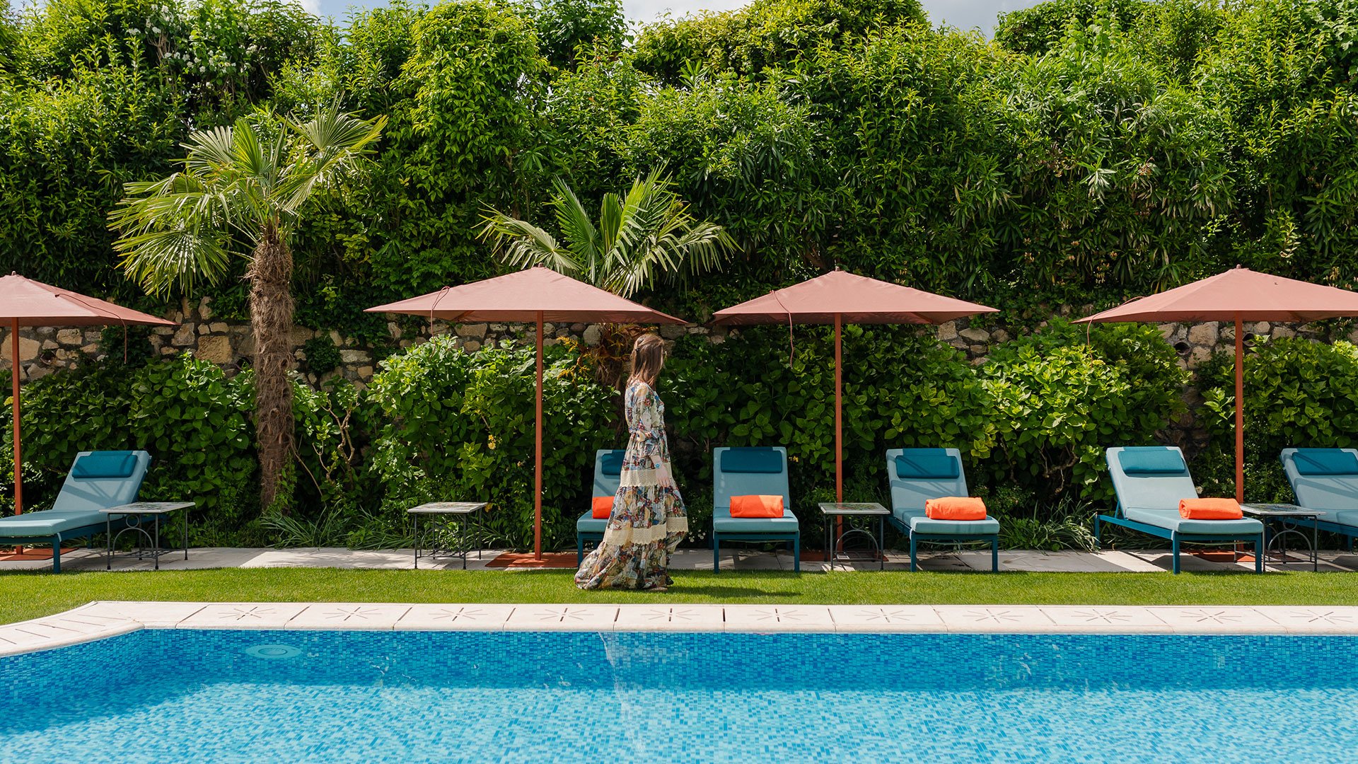 Pool area surrounded by greenery with blue sunbeds, umbrellas and orange towels at Byblos Art Hotel